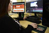 Mulher está sentada em uma mesa, usando um teclado e mouse enquanto trabalha em um computador com dois monitores. O monitor da esquerda exibe a página web de EAD da Unoeste, enquanto o monitor da direita mostra um software de edição de vídeo. Ela está usando uma blusa preta e branca e tem um copo da Unoeste na mesa ao lado do monitor. 