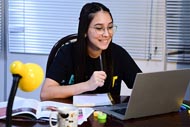 Uma jovem com óculos e camiseta preta está sentada à mesa, concentrada em seu laptop. Ela segura uma caneta na mão direita, enquanto um caderno aberto está à sua frente. A mesa de madeira escura também contém uma caneca, um livro aberto, rolos de fita adesiva colorida e algumas canetas. Uma luminária amarela está posicionada ao fundo, perto da janela com persianas. A cena sugere um ambiente de estudo, com a jovem possivelmente assistindo a uma aula online, fazendo anotações ou realizando alguma tarefa no computador.