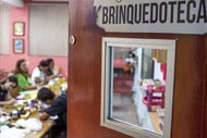 A imagem mostra a entrada de uma brinquedoteca, com uma porta parcialmente aberta que revela um ambiente interno onde várias pessoas estão envolvidas em atividades criativas. A sala é decorada com paredes coloridas e mesas repletas de materiais artísticos, como tintas e pincéis. O espaço parece ser voltado para o desenvolvimento lúdico e educativo para crianças.